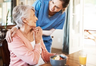 Seniorin beim Frühstück mit Pflegerin im Hintergrund